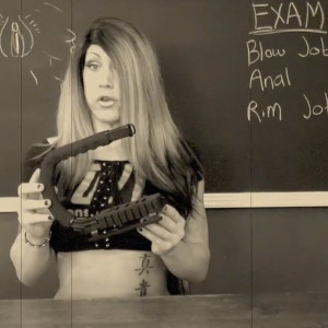 Kimber Haven is pictured in a retro style black-and-white photo at a blackboard, holding a strange sex toy.