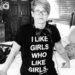 Black and white photo of short-haired bespectacled Mills in a black t-shirt that reads I Like Girls Who Like Girls.